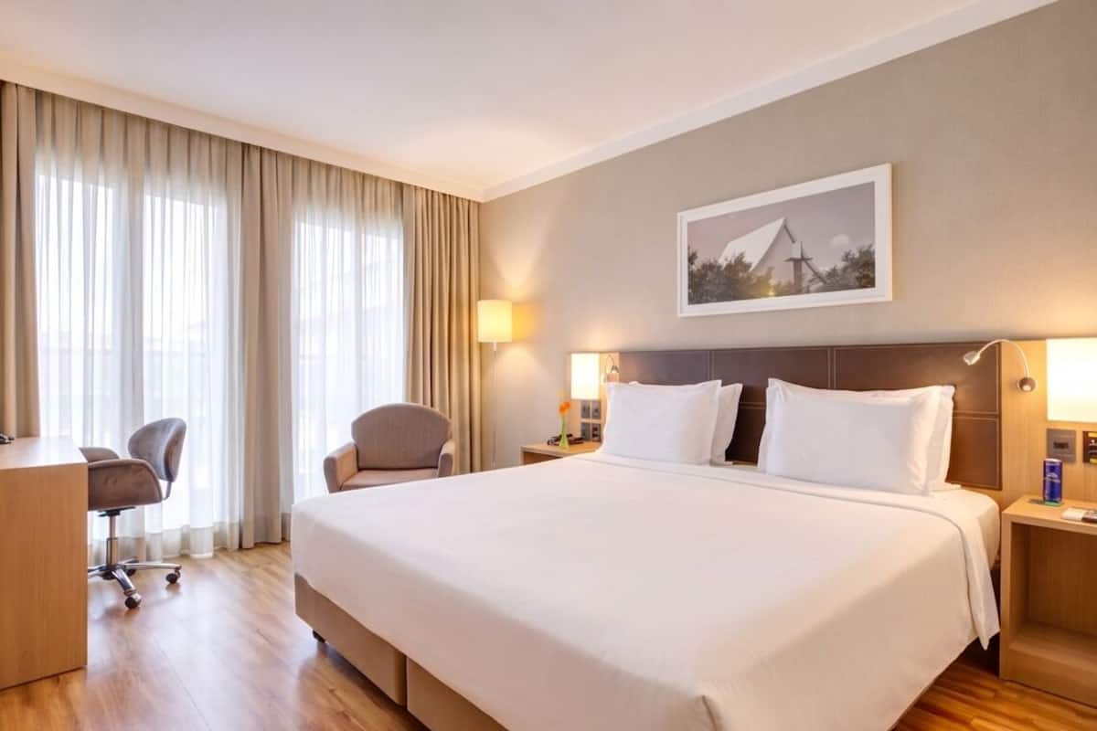 A hotel room with a large bed, two bedside tables, a desk, a chair, and a large window with curtains.