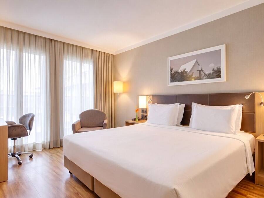 Quarto de hotel com uma cama grande, duas mesas de cabeceira, uma escrivaninha, uma cadeira e uma janela ampla com cortinas.