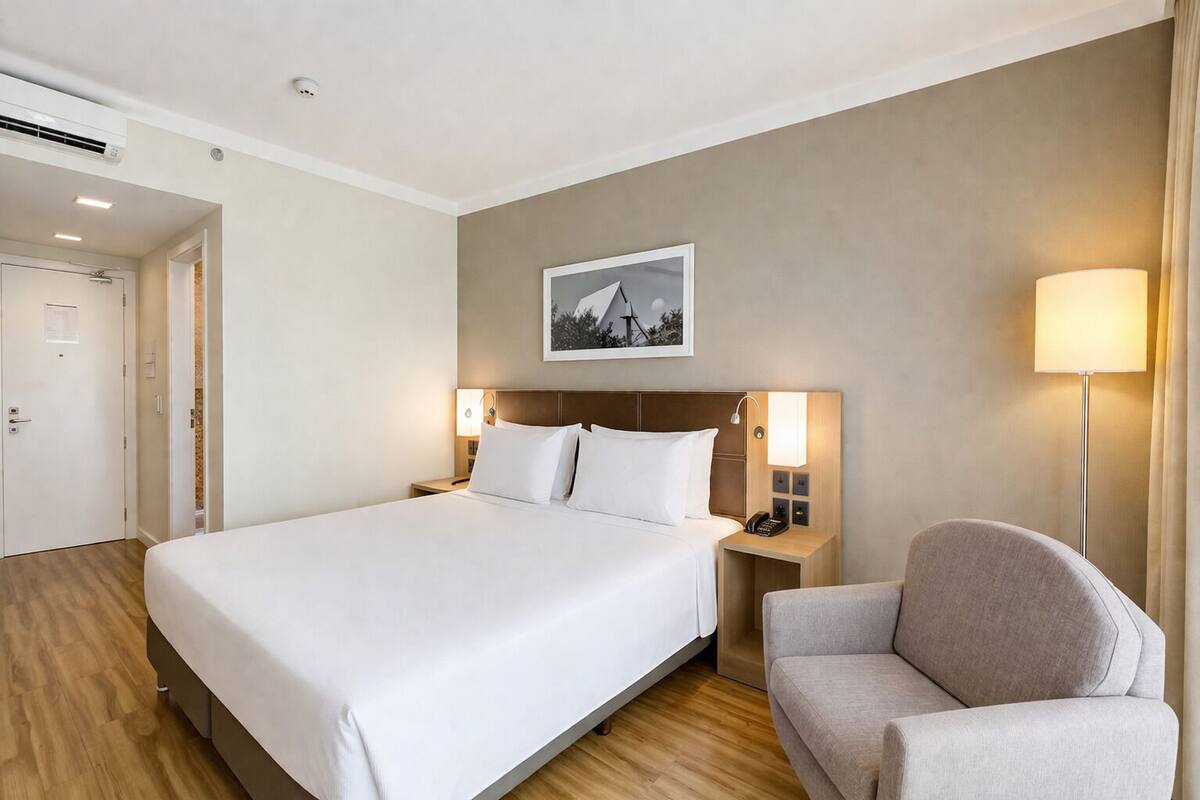 A hotel room with a large bed, bedside tables with lamps, a chair, a framed picture on the wall, and an air conditioning unit.