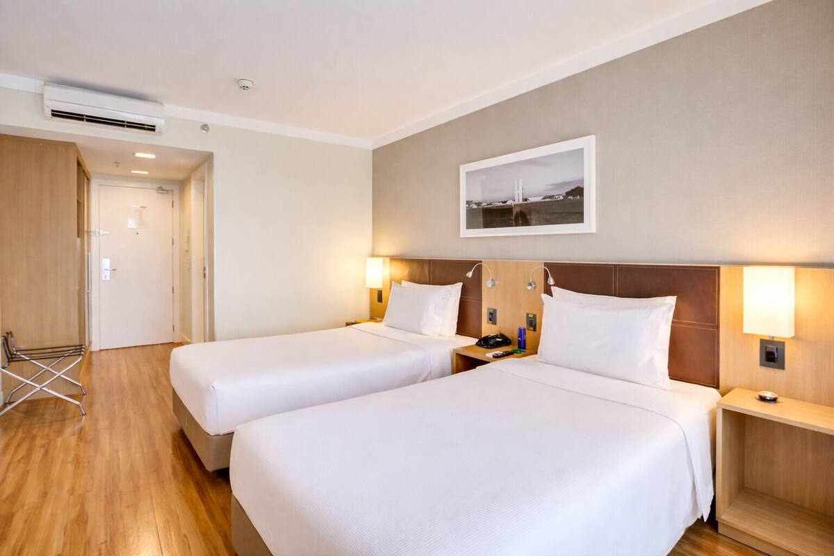 A hotel room with two beds, a wooden floor, a wall-mounted air conditioner, a door, and a framed picture on the wall.