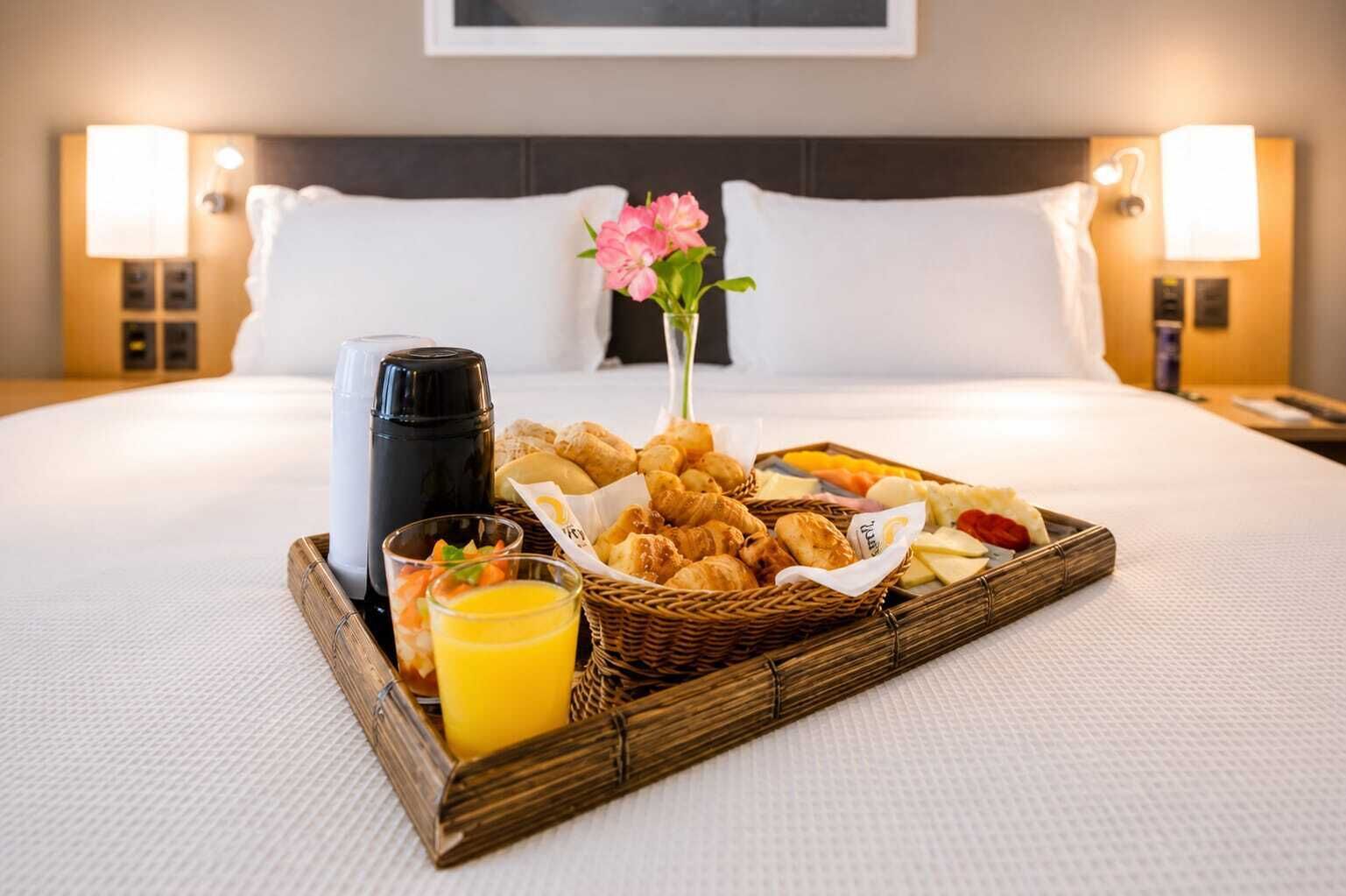 A neatly made breakfast tray on a hotel bed with croissants, pastries, fruits, and a glass of orange juice.