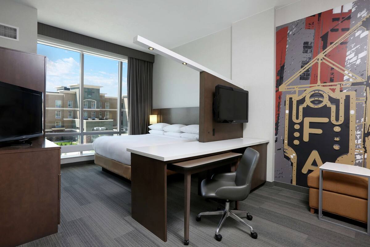 A hotel room with a large bed, a desk with a chair, a TV, and a large window offering a city view.