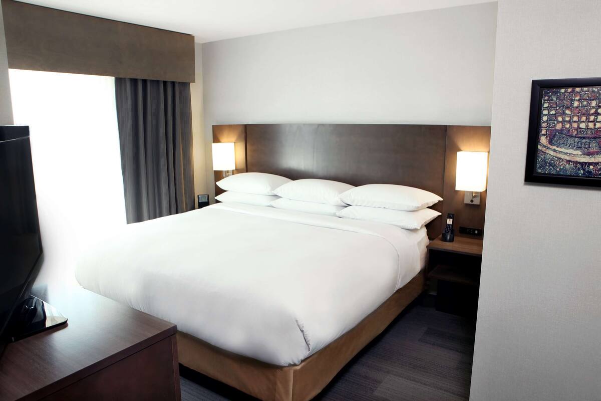 A hotel room with a large bed, two bedside lamps, a TV on a wooden nightstand, and a framed artwork on the wall.