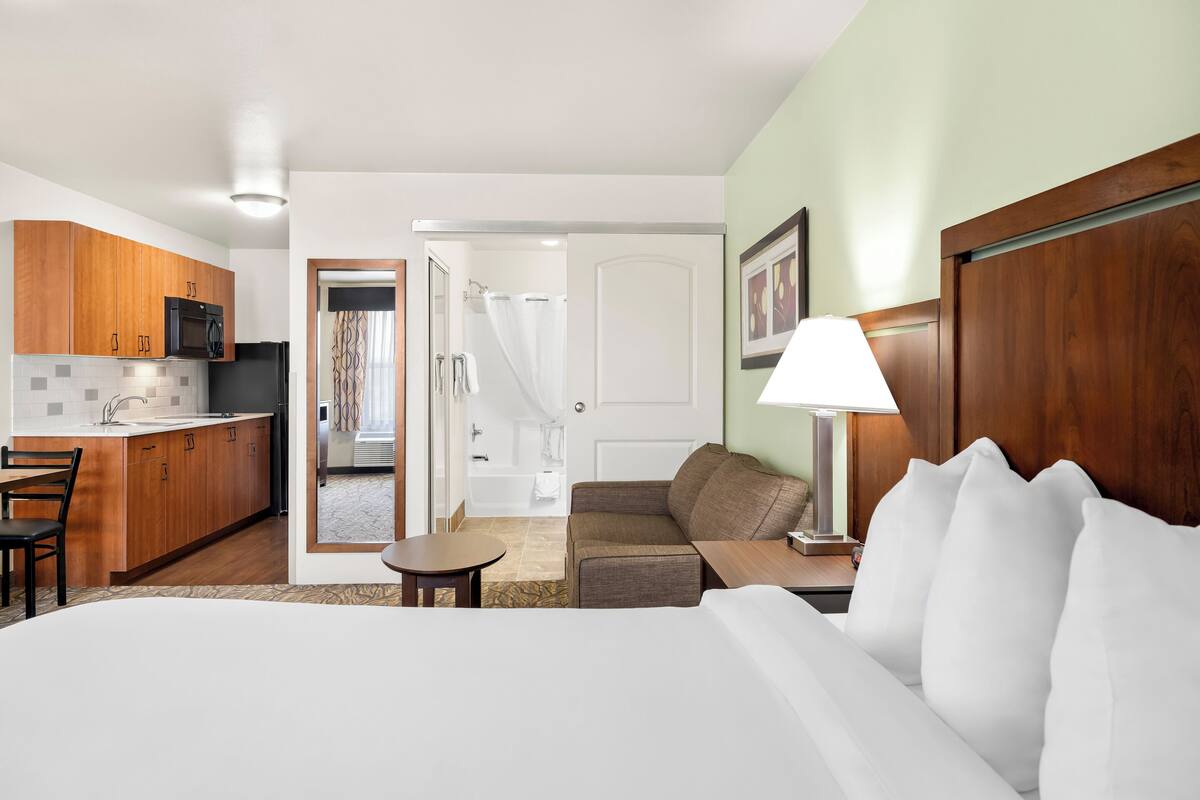 A hotel room with a bed, a desk, a chair, a small table, a lamp, a sofa, a bathroom visible through an open door, and wooden cabinets.