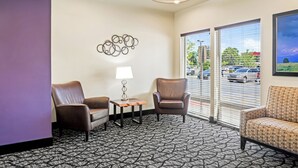 A hotel room with a patterned carpet, two leather chairs, a small table with a lamp, a wall art piece, and large windows with blinds. - Ledgestone Suites Hotel Billings (Billings)