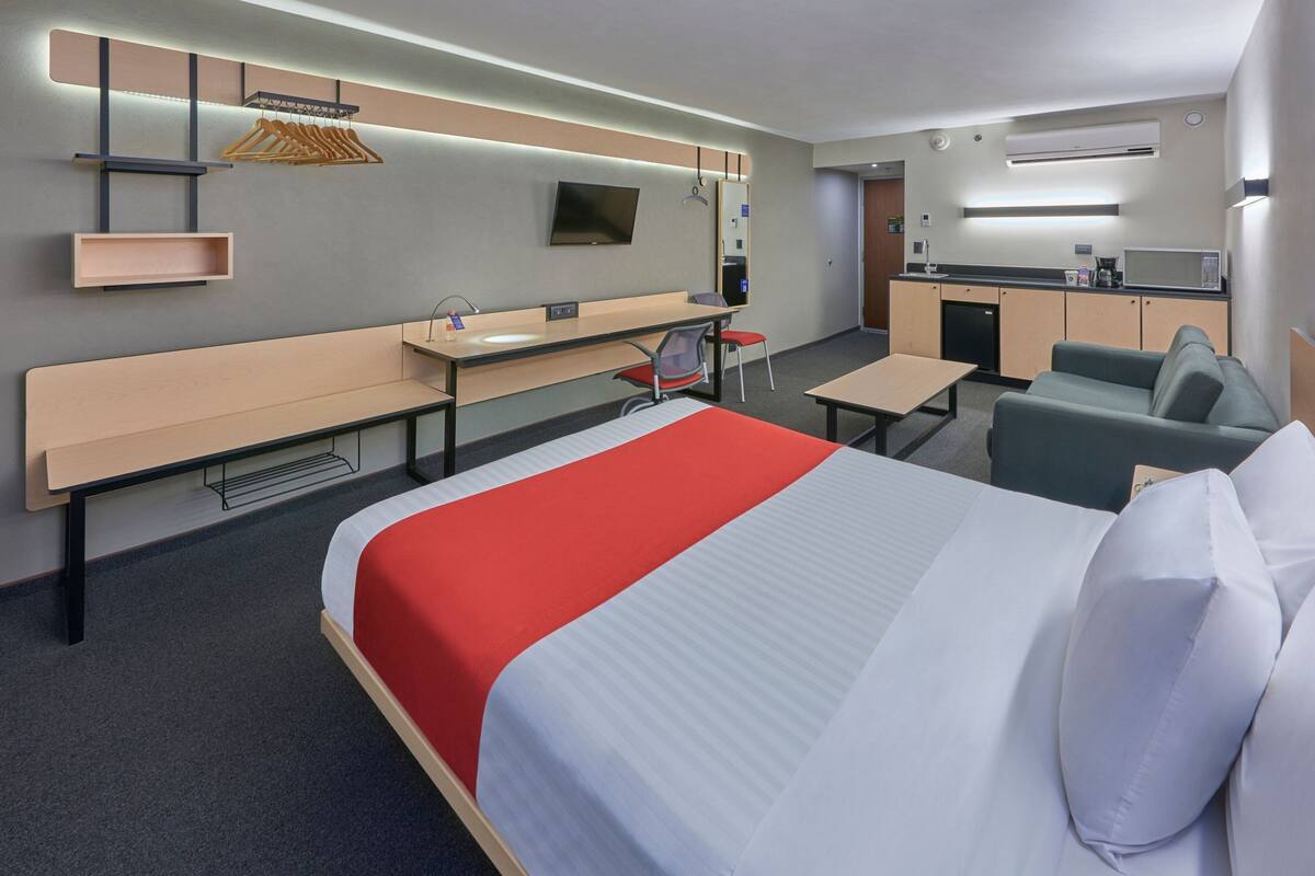 A hotel room with a bed, a desk, a chair, a sofa, a TV, and a kitchenette.