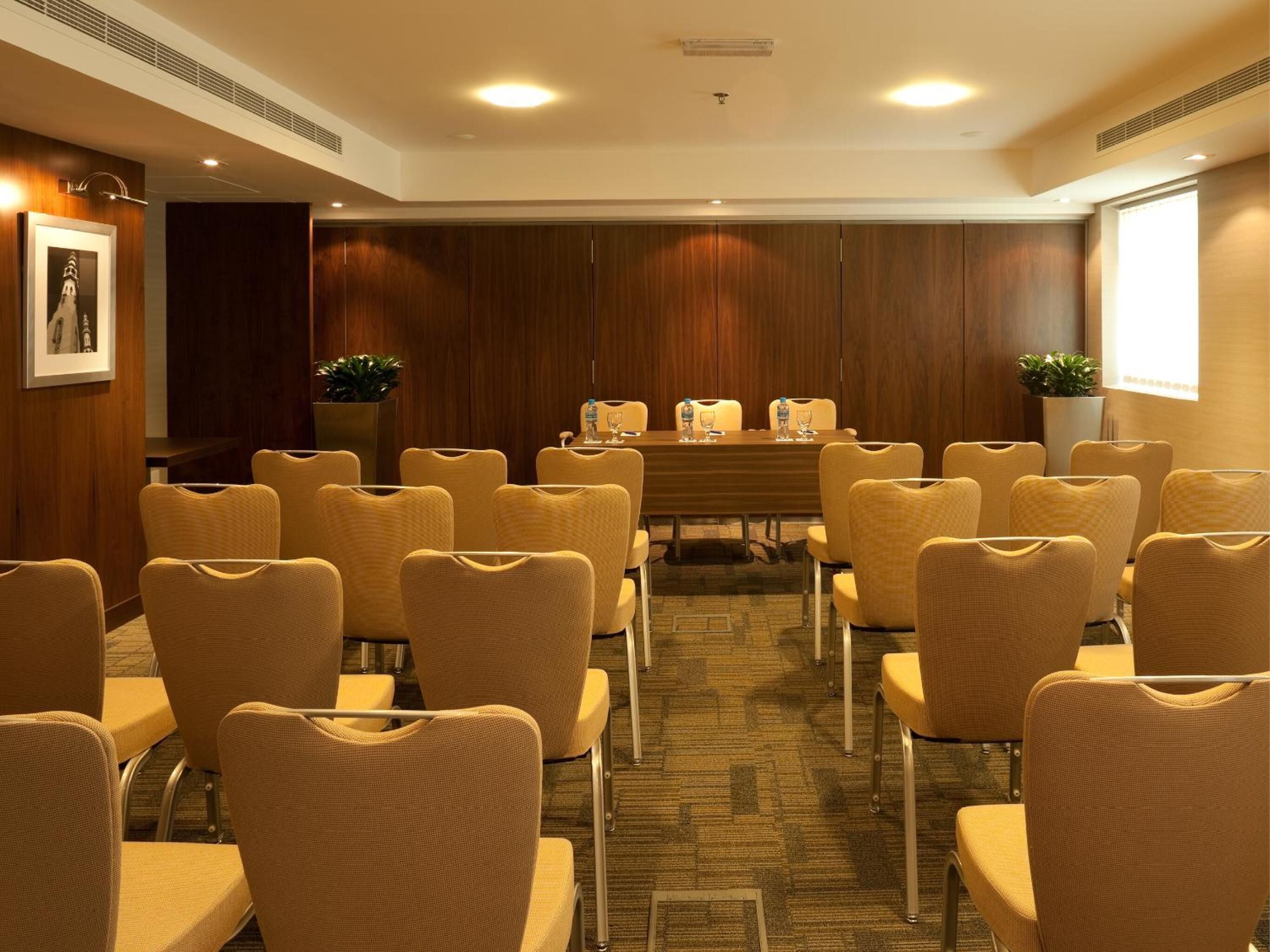 A conference room with a long table and multiple chairs, wooden panel walls, and two potted plants.