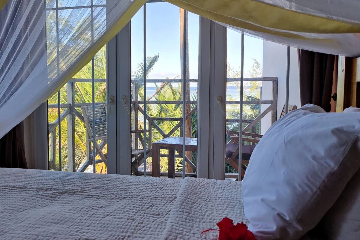 A bed with a white quilt and a red accent pillow, a wooden headboard, and sheer curtains.