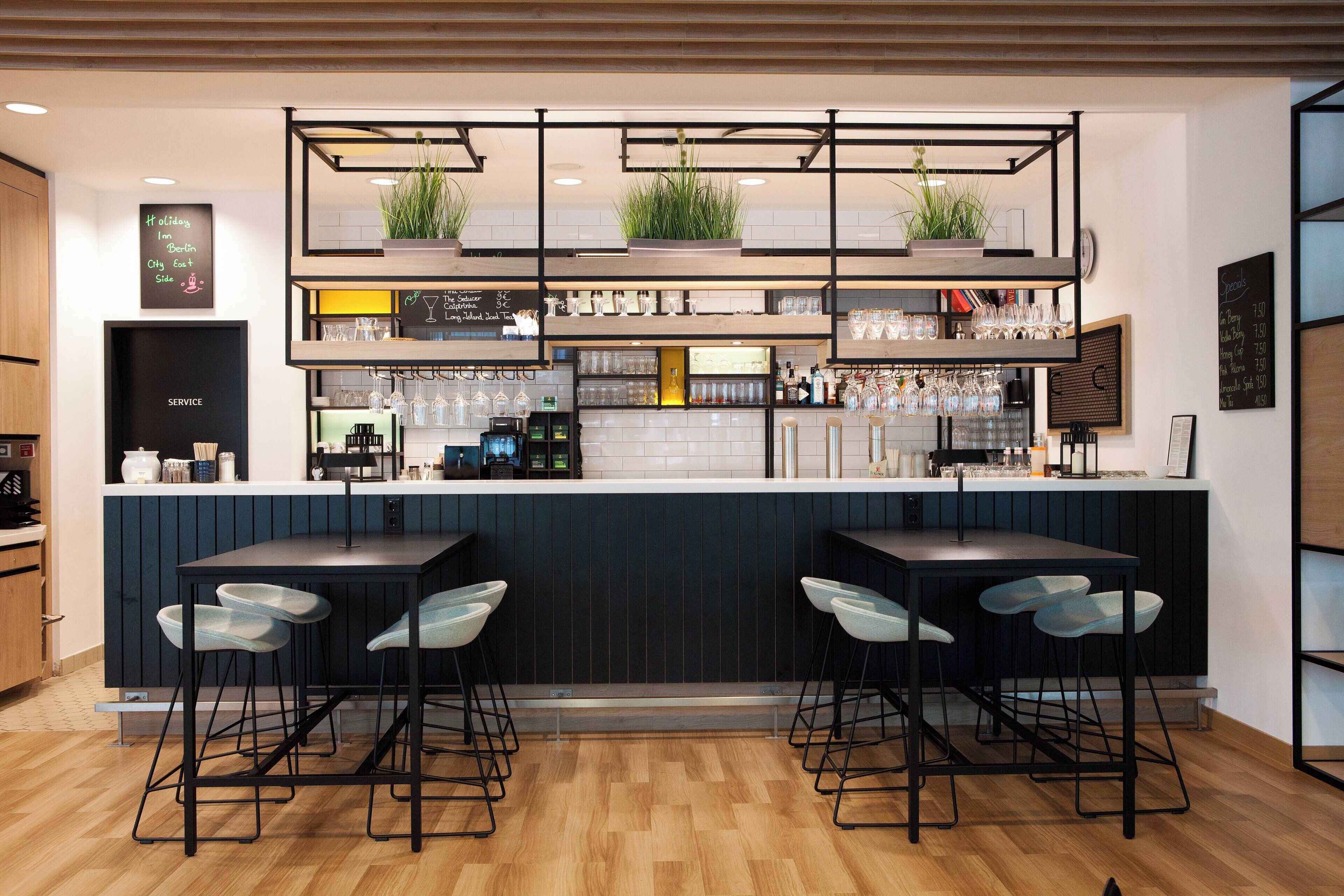 Zona de bar con mostrador, taburetes y estantes con copas y plantas.