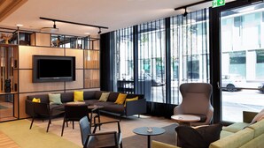 A modern lounge area with a flat-screen TV mounted on the wall, comfortable seating including sofas and chairs, and large windows offering a view of the street. - Holiday Inn Berlin - City East Side by IHG (Berlin)