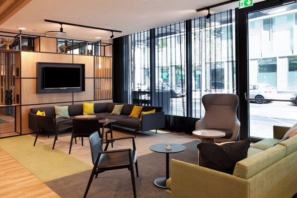 A modern lounge area with a flat-screen TV mounted on the wall, comfortable seating including sofas and chairs, and large windows offering a view of the street. - Holiday Inn Berlin - City East Side by IHG (Berlin)