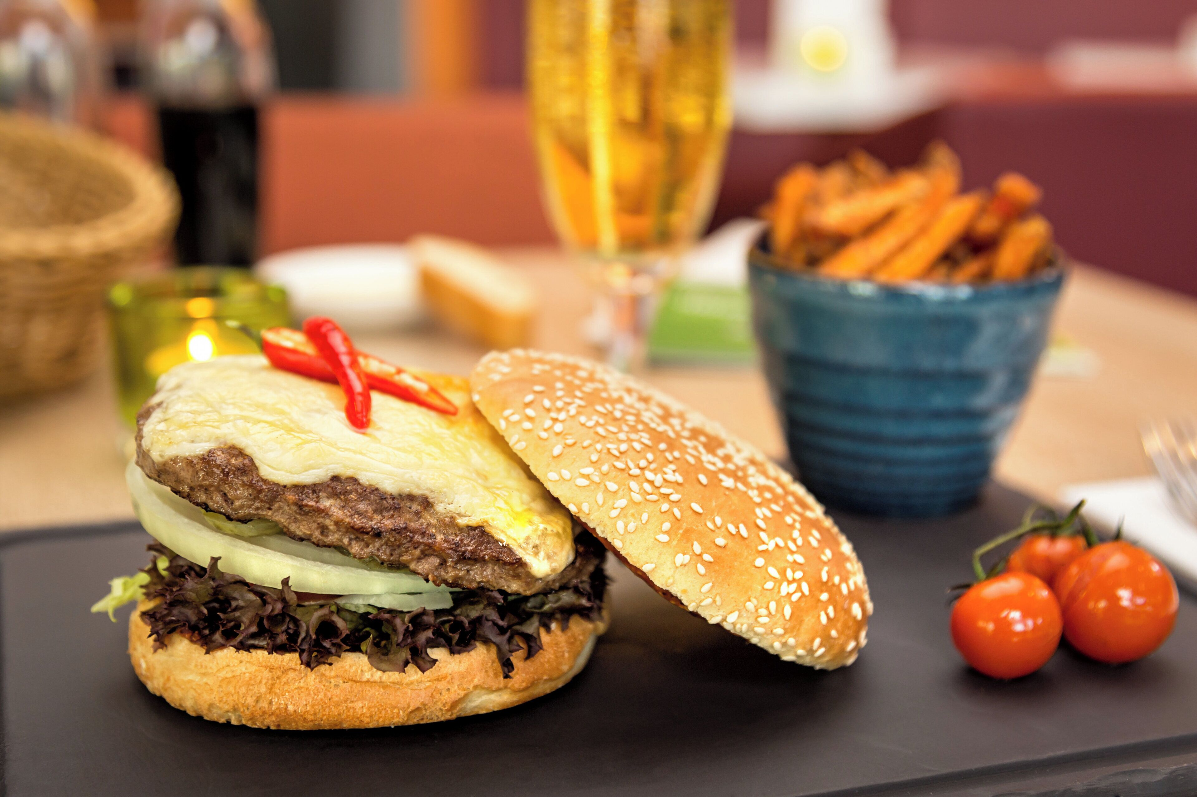 A burger with lettuce, tomato, and a red chili pepper on top, accompanied by a side of fries and a glass of a yellow beverage.