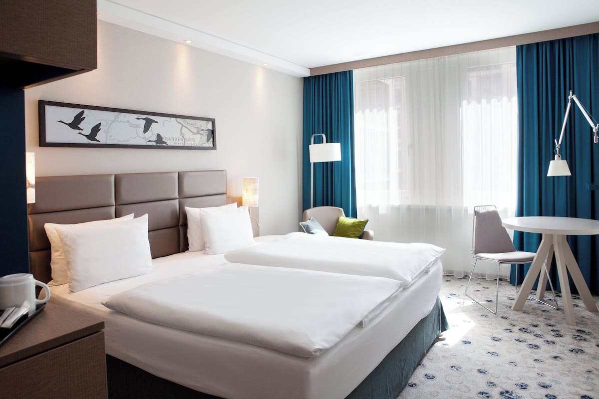 Habitación de hotel con dos camas, un escritorio, una silla, una mesa y un gran ventanal con cortinas.
