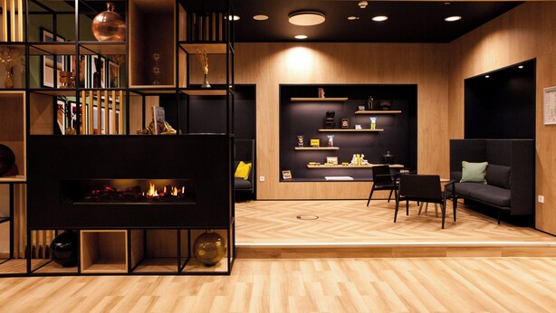 A modern living room with a fireplace, wooden flooring, and a black sectional sofa. - Holiday Inn Berlin - City East Side by IHG (Berlin)