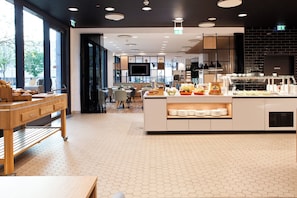 Una cafetería moderna con un suelo de baldosas hexagonales, una barra central con diversas opciones de comida y un área de estar con mesas y sillas.