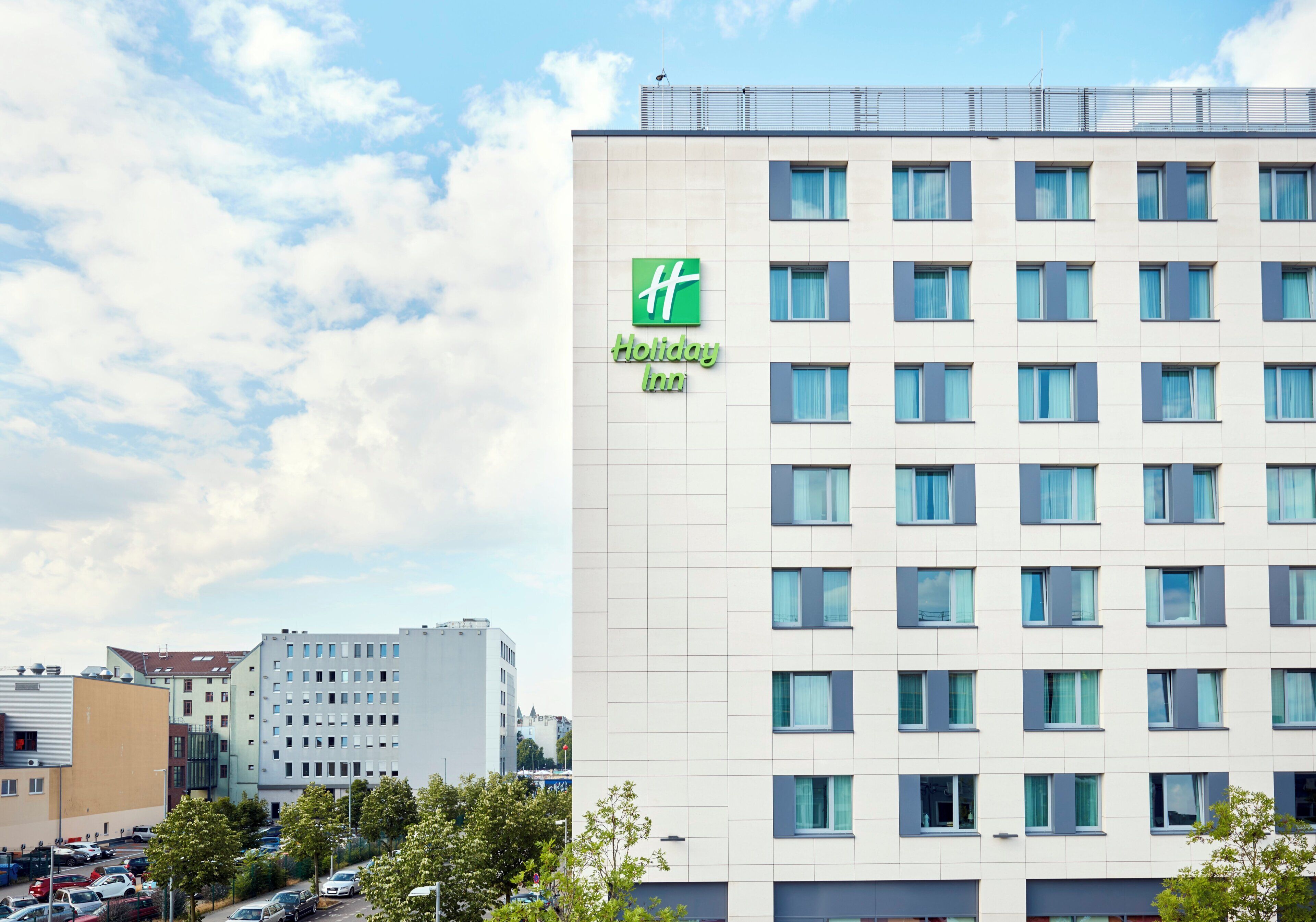 Un hotel de varios pisos con un logotipo verde llamativo, una fachada moderna y un estacionamiento al frente.