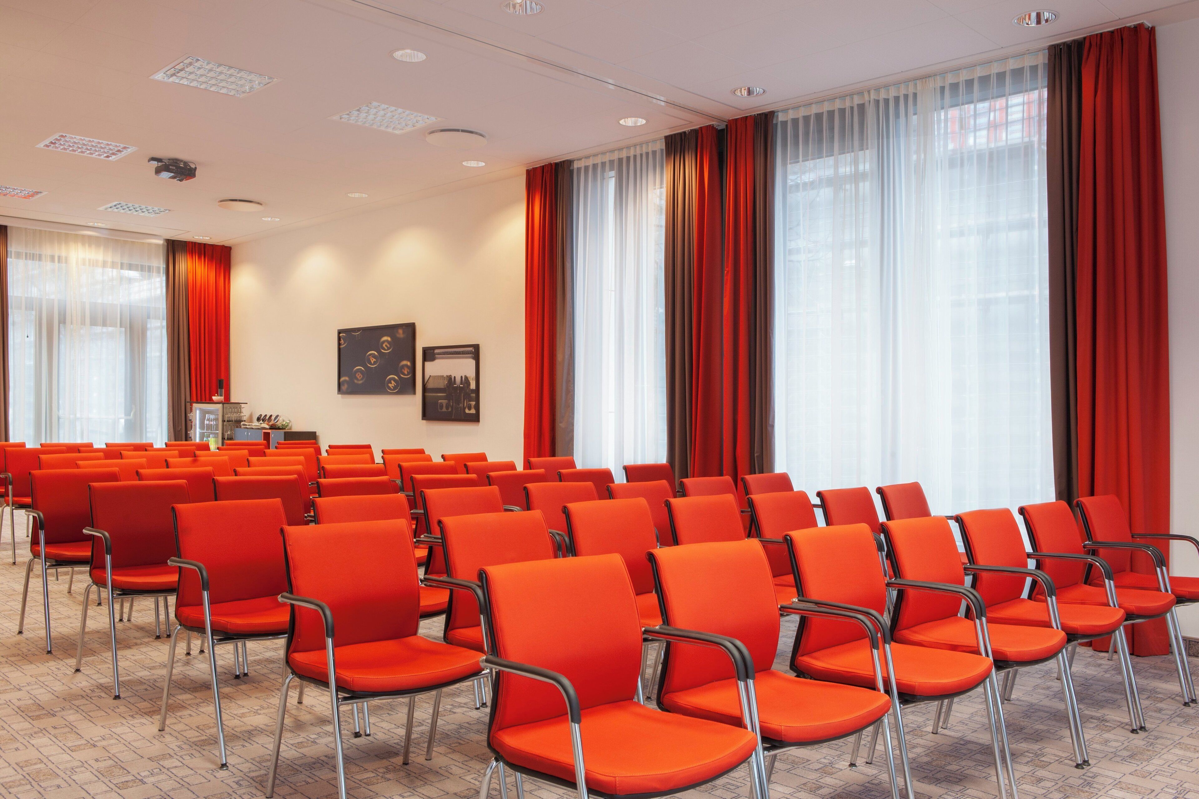 Un salón de conferencias con filas de sillas anaranjadas, ventanas amplias con cortinas traslúcidas y una zona de kitchenette.