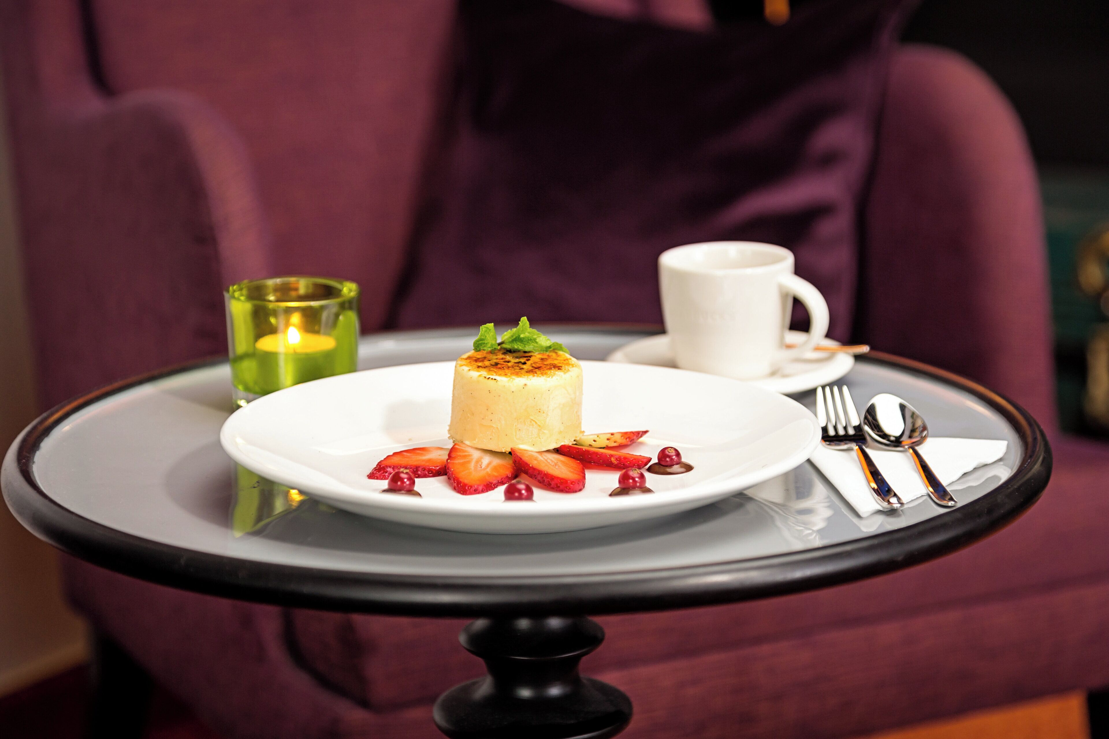 A dessert on a white plate with strawberries and a candle on a table.