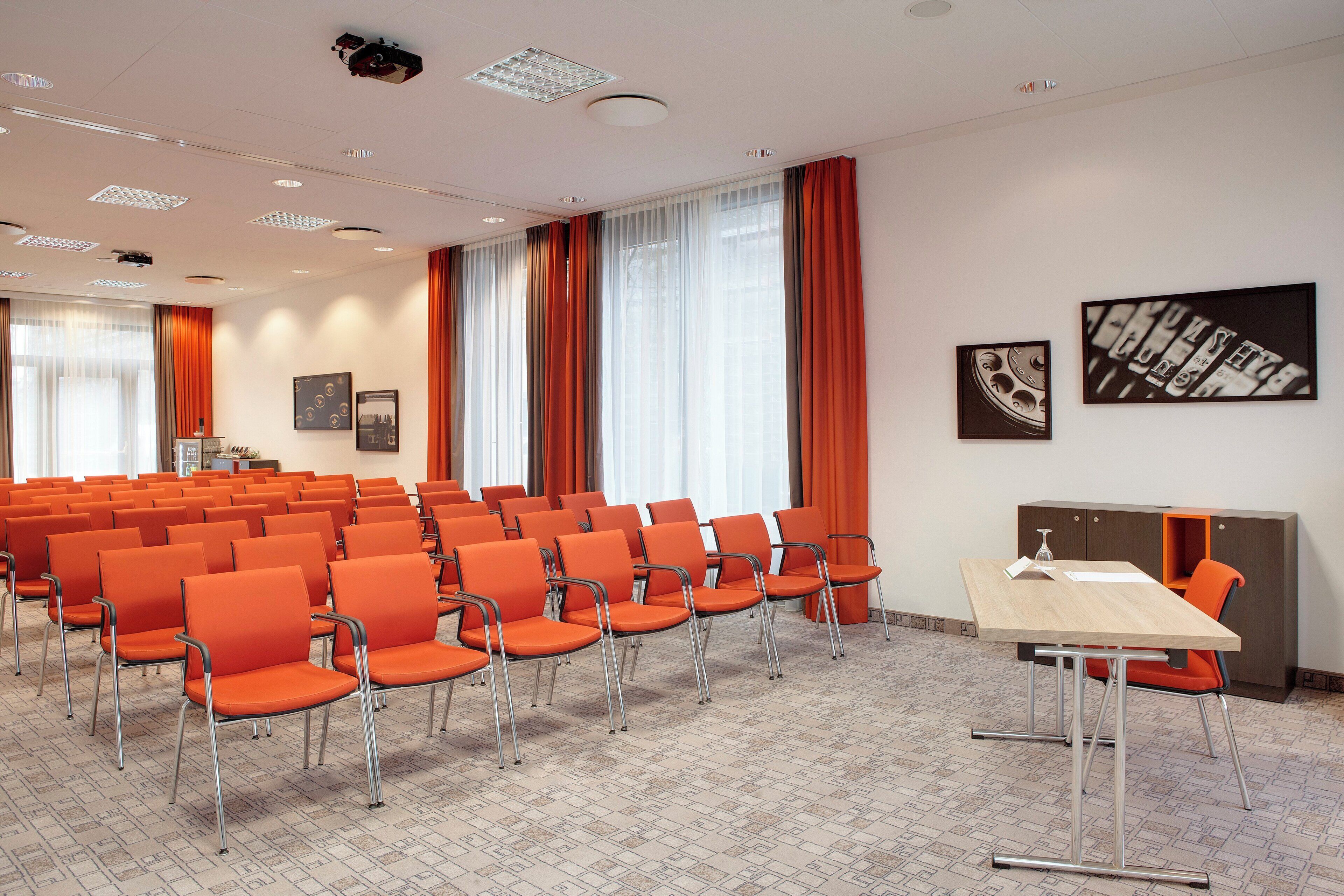 Une salle de conférence avec des rangées de chaises orange, une table en bois et de grandes fenêtres ornées de rideaux orange.