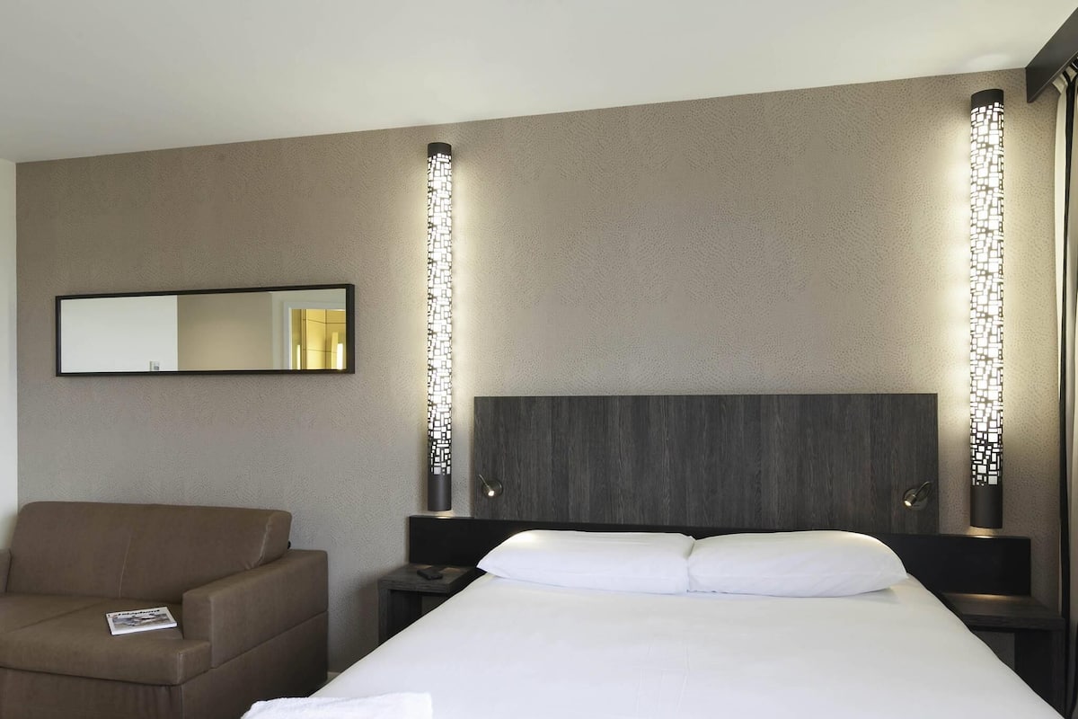 Ein Hotelzimmer mit Bett, Stuhl, Nachttisch, Spiegel und Wandleuchten.