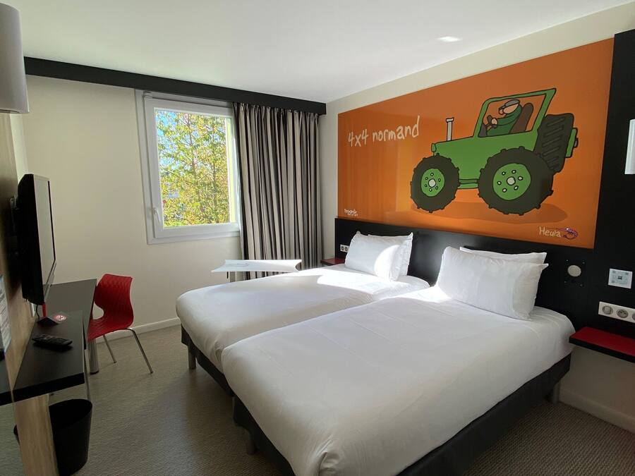 A hotel room with two beds, a desk with a chair, a TV, a window with curtains, and a wall mural of a green tractor.