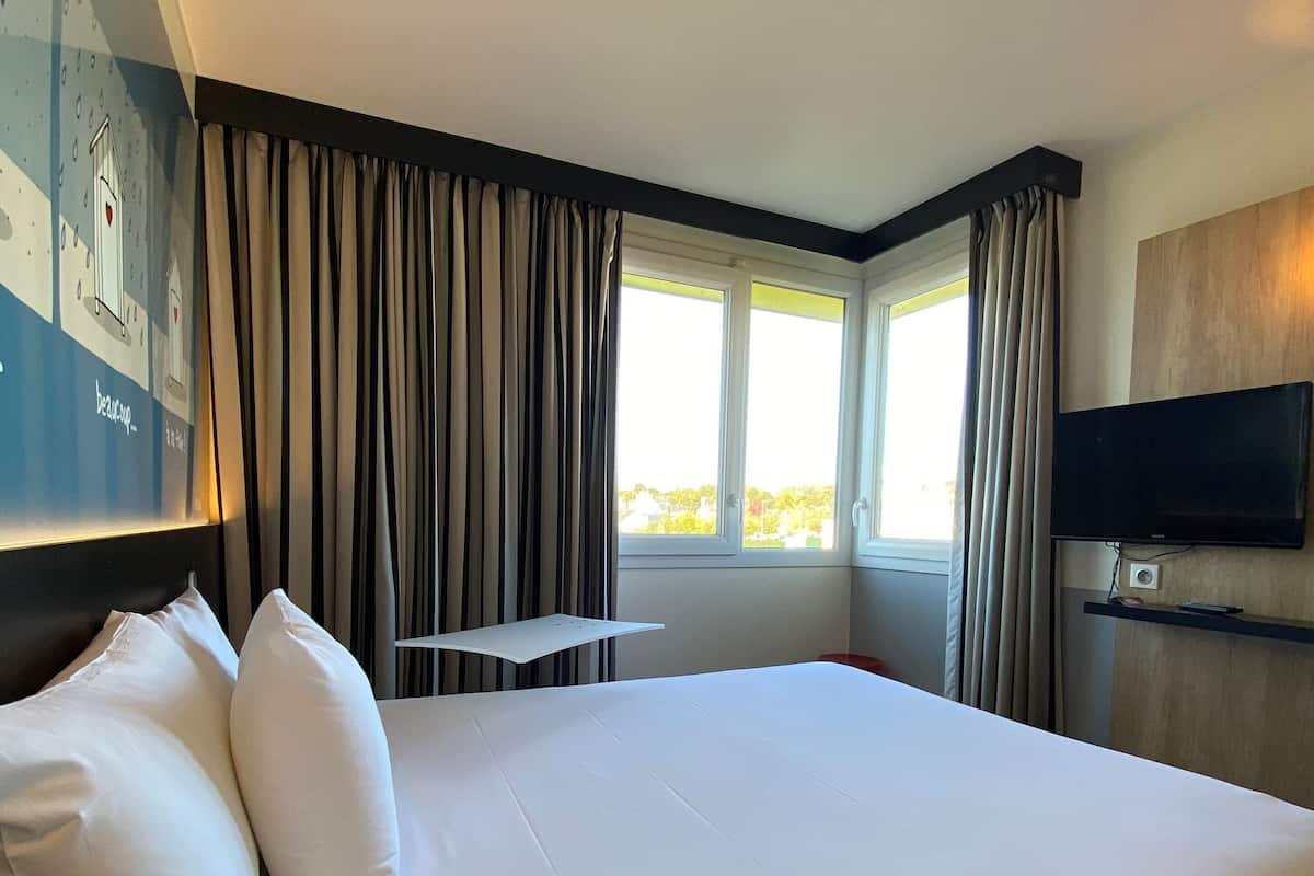 A hotel room with a large bed, a flat-screen TV, a small table, and a window with curtains.