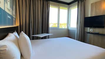 A hotel room with a large bed, a flat-screen TV, a small table, and a window with curtains.