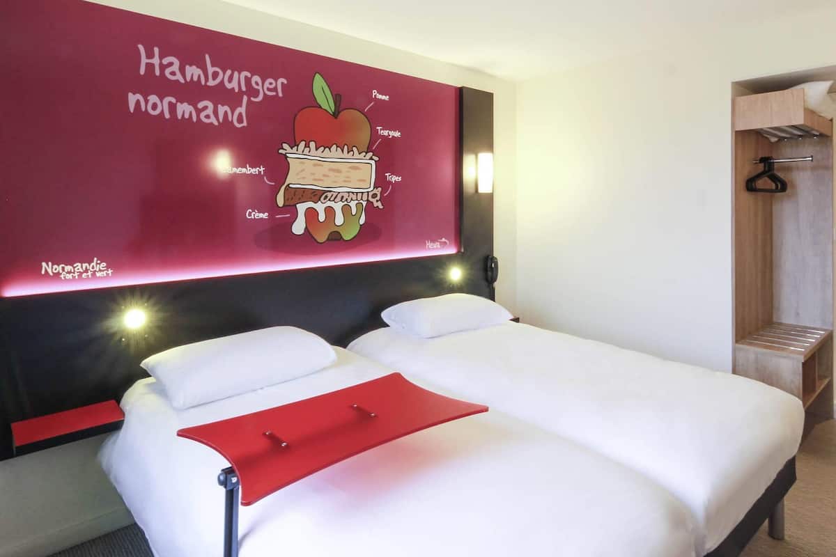 A hotel room with a large wall mural of a hamburger, a bed with white linens, and a red bedside table.