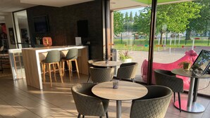 A modern cafe with round tables and chairs, a bar area, and large windows offering a view of a park. - Ibis Styles Flers (Flers)