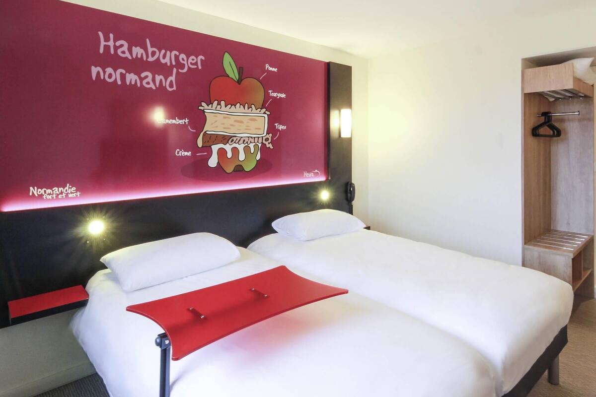 A hotel room with a bed, a red wall with a food illustration, a phone, and a wooden bench.