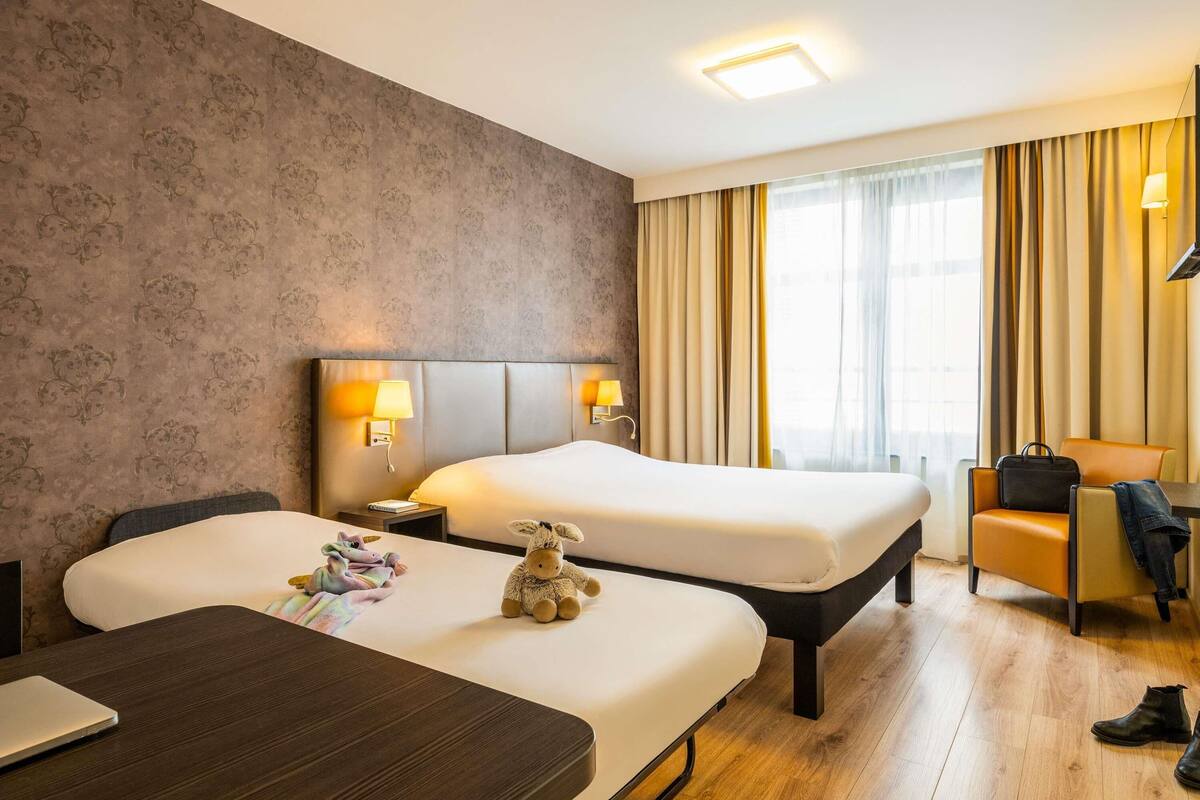 A hotel room with two beds, a desk, a chair, and a lamp.