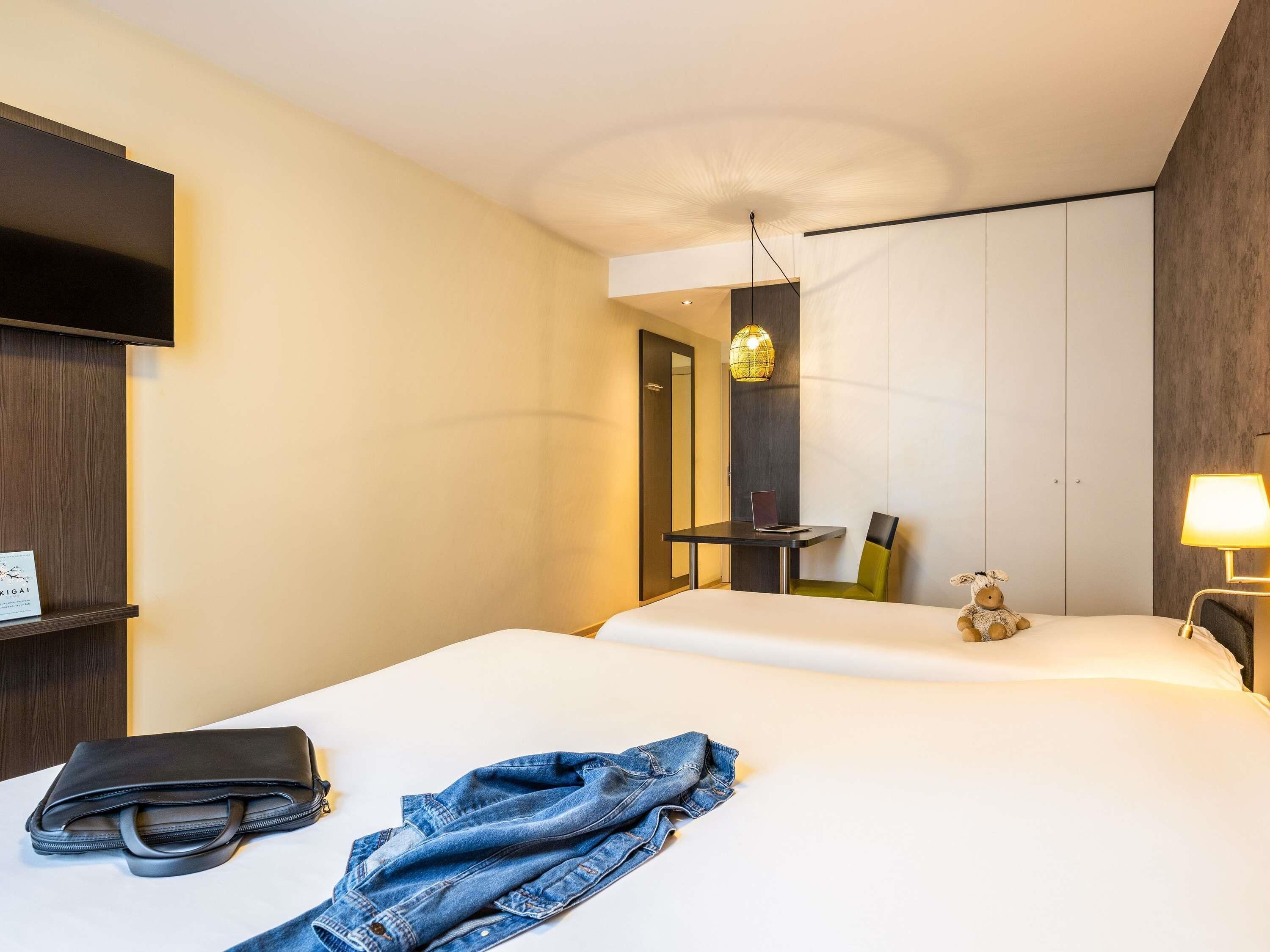 A hotel room with a bed, a desk with a laptop, a chair, a lamp, and a teddy bear.