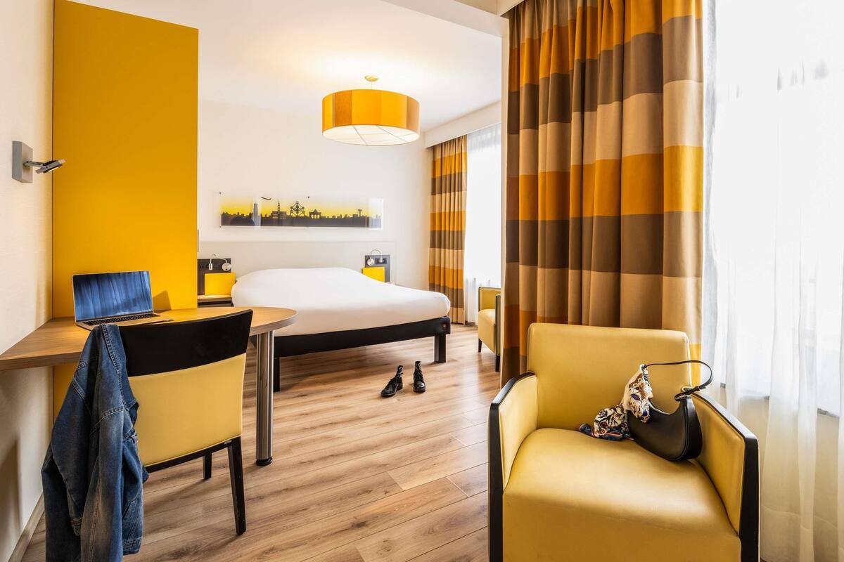 A hotel room with a bed, a desk, a chair, and a lamp.