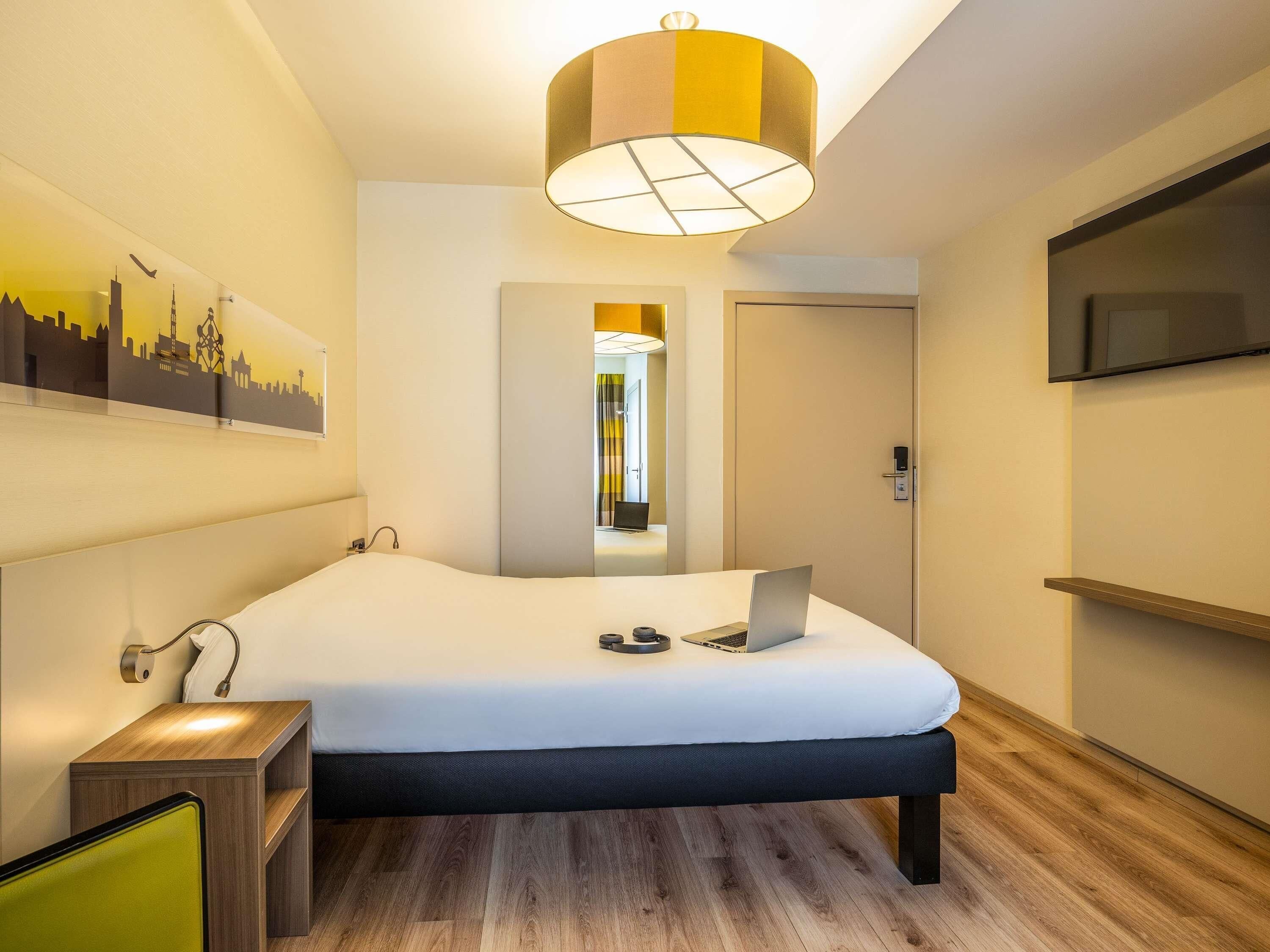 Habitación de hotel moderna con cama, escritorio, computadora portátil, televisión y una obra de arte con un paisaje urbano en la pared.