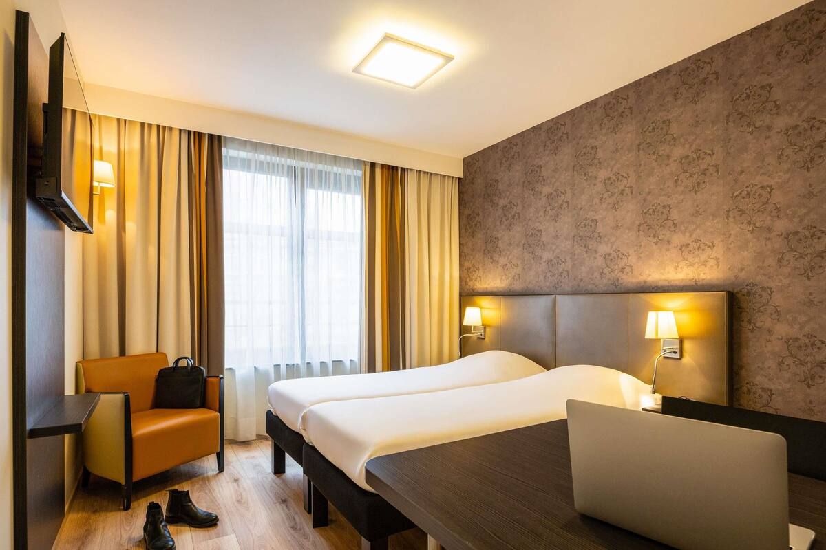 A hotel room with a bed, a desk, a chair, a laptop, and a pair of shoes on the floor.