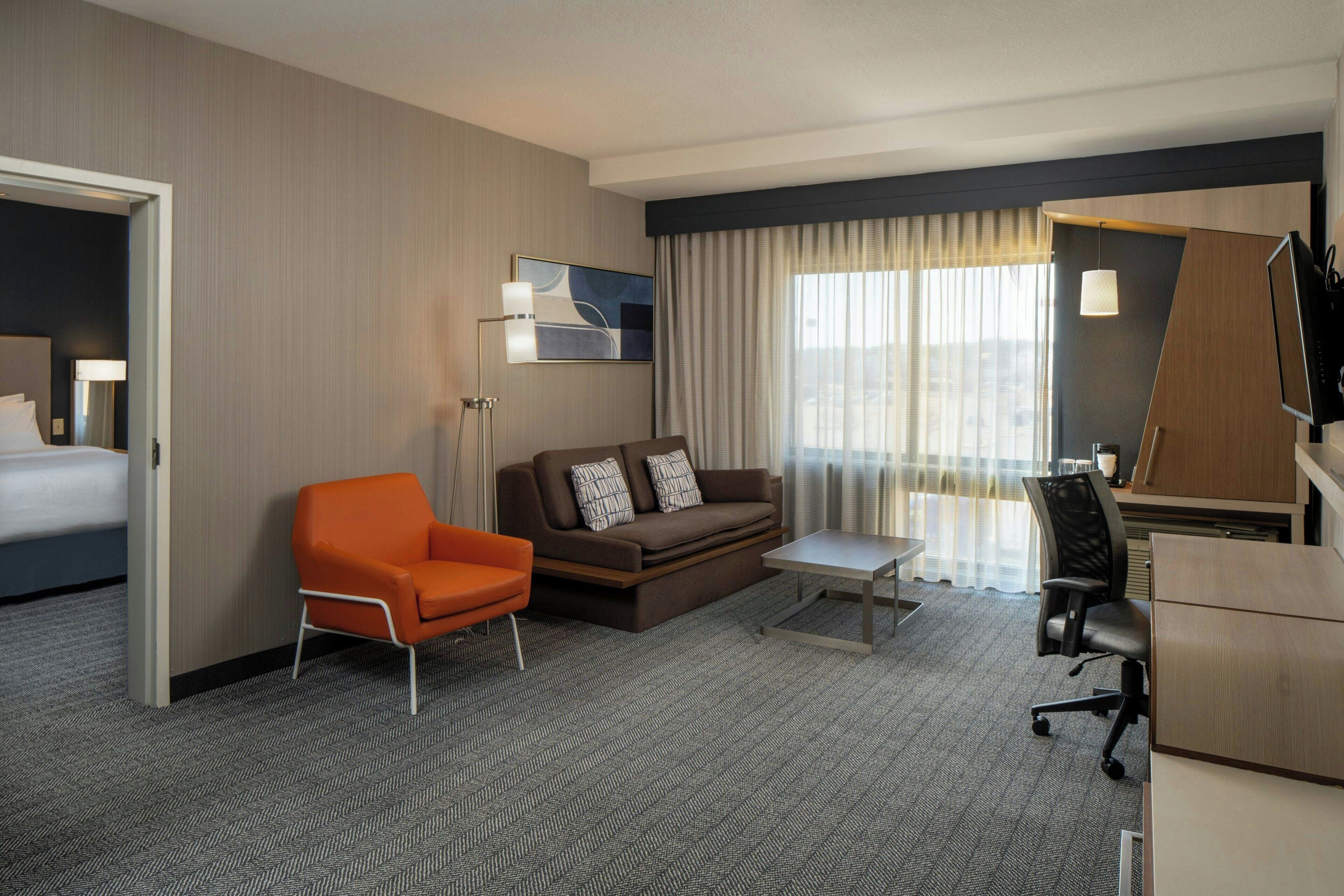 Una habitación de hotel con una cama, un sillón naranja, un sofá marrón, un escritorio con silla y un televisor.