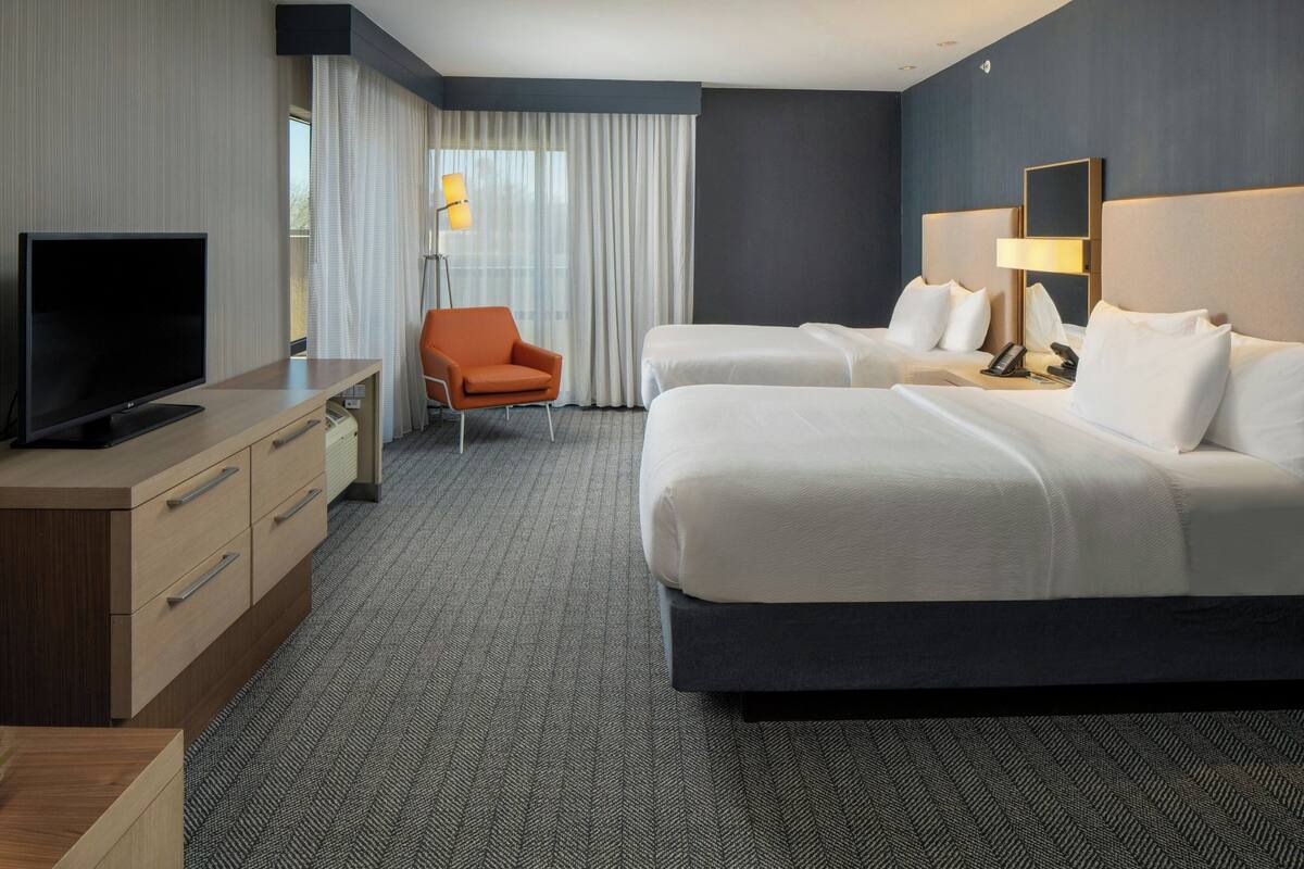 A hotel room with two beds, a desk with a TV, an orange chair, and a large window with curtains.