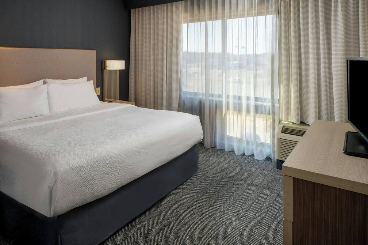 A hotel room with a large bed, a desk with a television, a lamp, and a window with curtains.