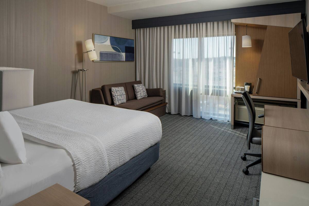 A hotel room with a bed, a desk, a chair, a lamp, and a large window with curtains.