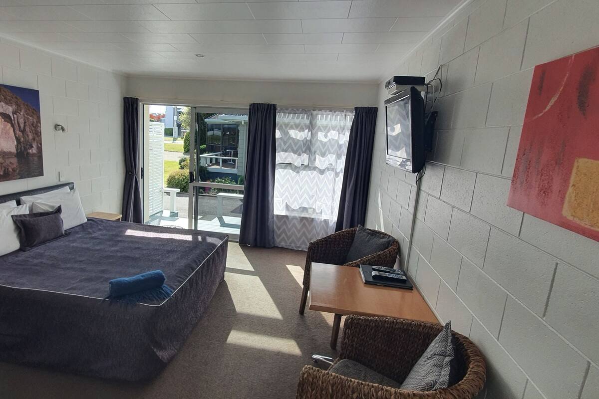 Habitación de hotel con cama, una mesita, una silla, televisor y un cuadro en la pared.
