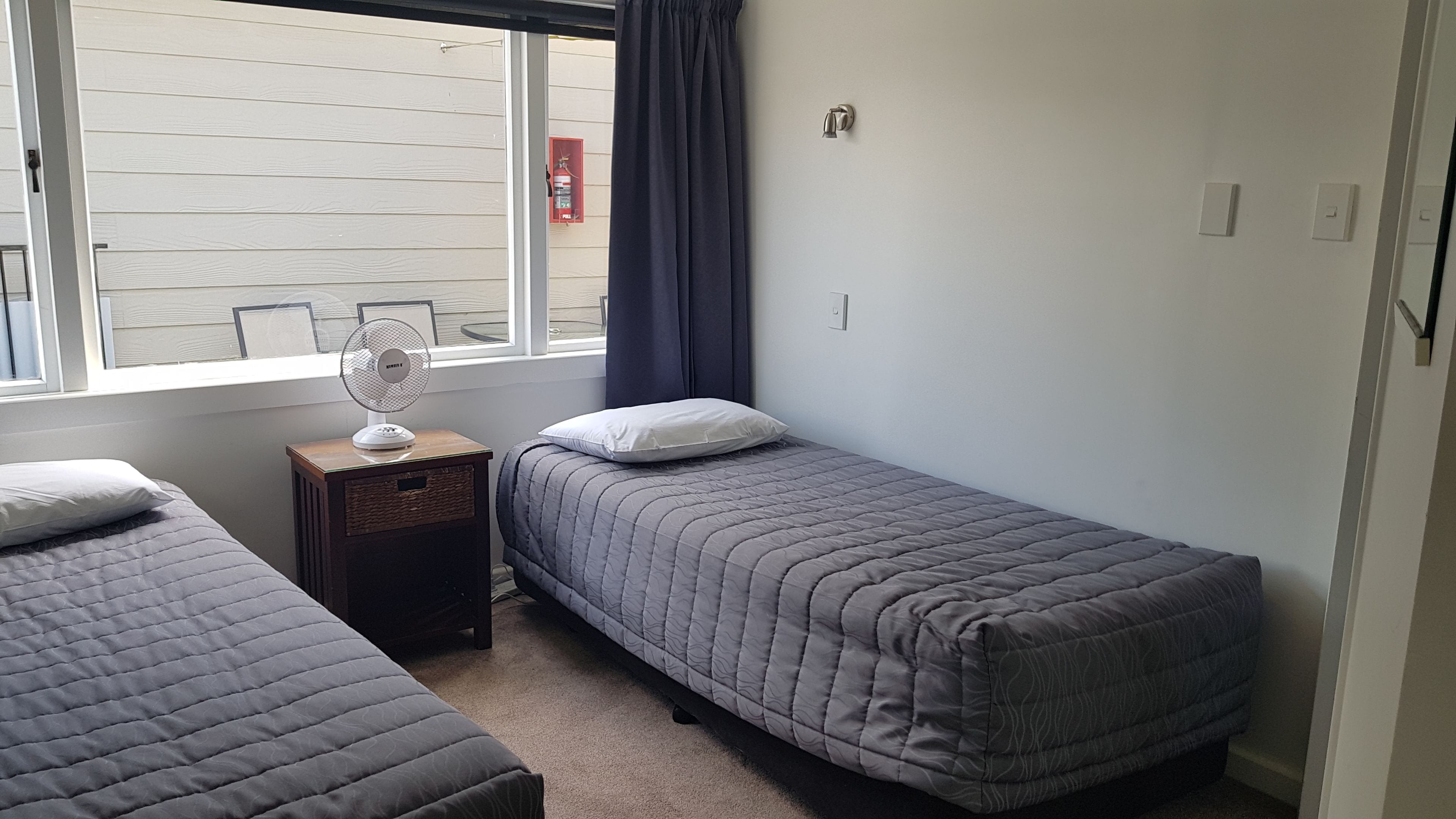 Una habitación con dos camas individuales, un ventilador de pie, una ventana con cortinas y un extintor.