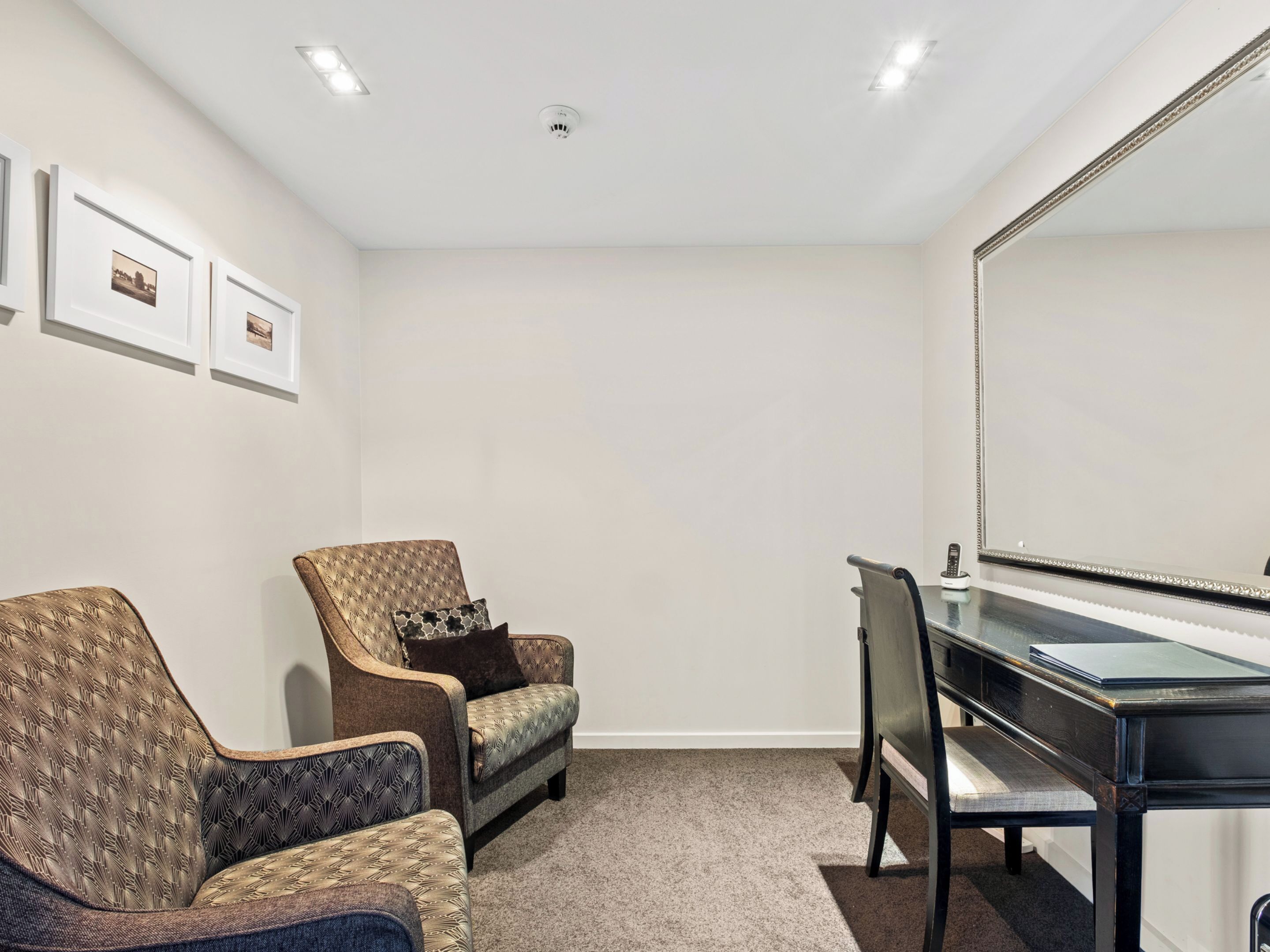 Una habitación con un espejo grande, dos sillones, una mesita y suelo alfombrado.