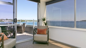 Een balkon met een stoel en een bank, grote ramen met uitzicht op zee en bergen, en een plant in een pot.