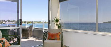 A balcony with a chair and a sofa, large windows with a view of the sea and mountains, and a potted plant.