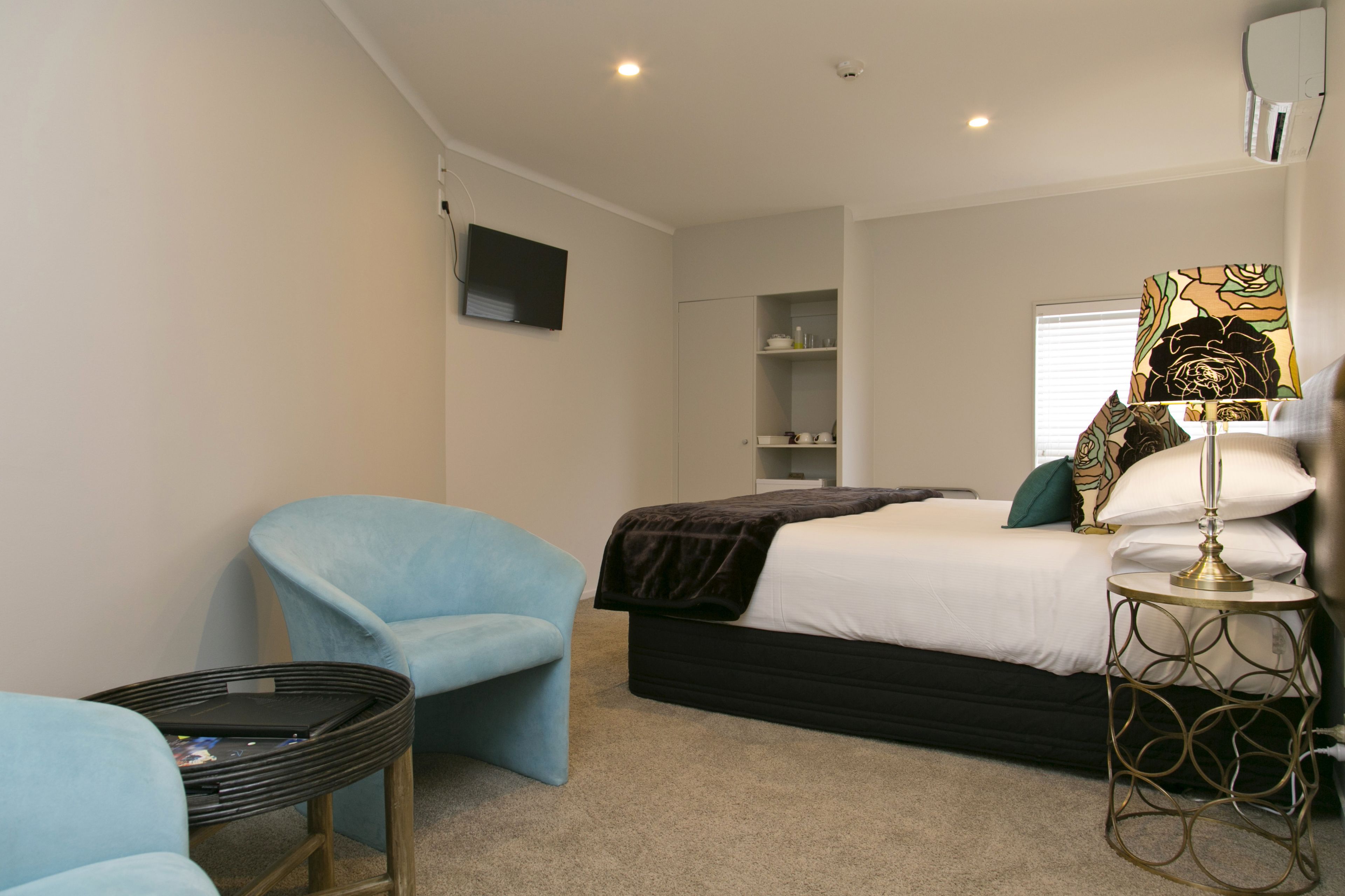 A bedroom with a bed, a blue armchair, a bedside table with a lamp, and a TV mounted on the wall.