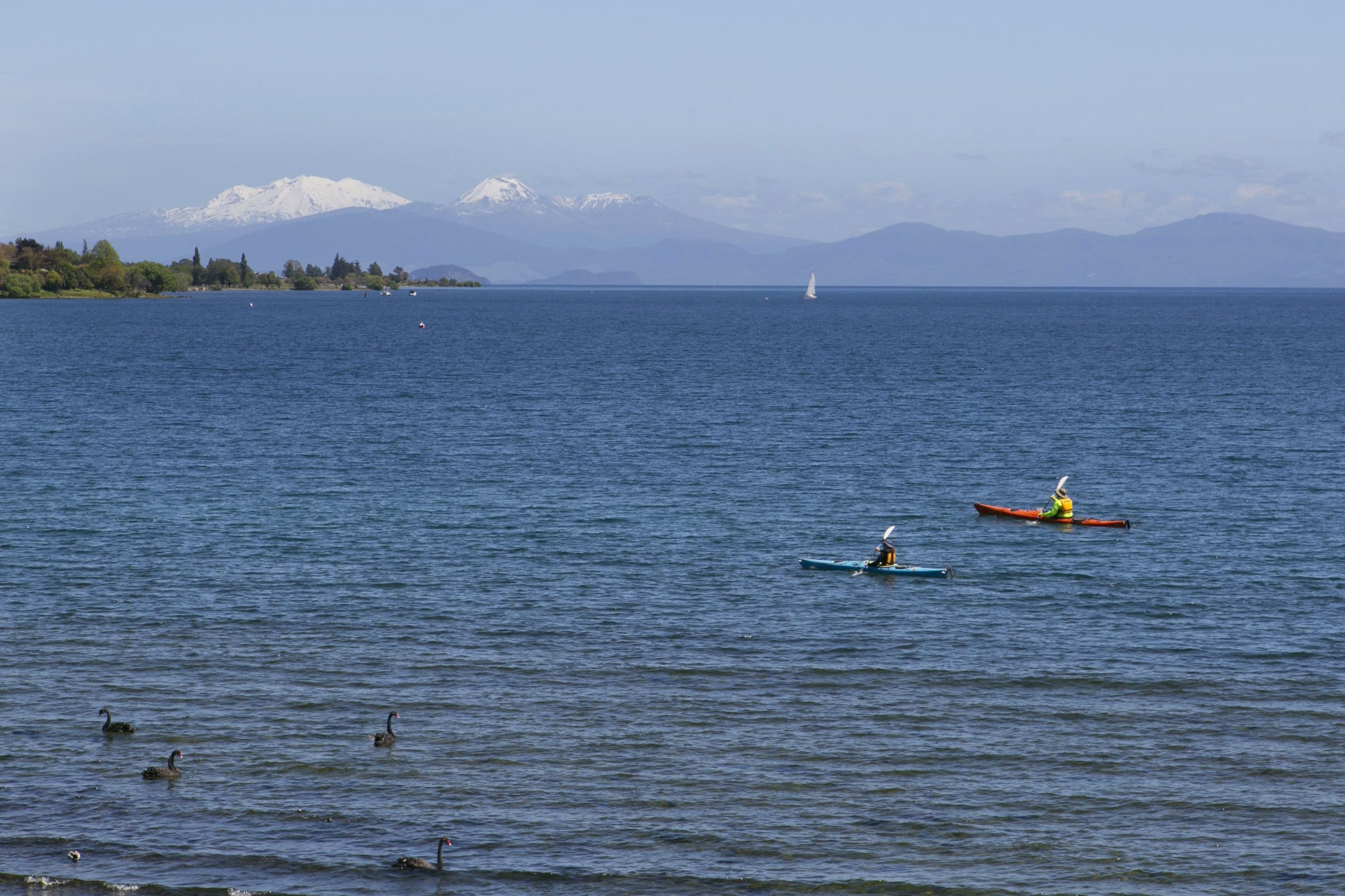 A calm lake with kayaks and a sailboat, surrounded by mountains and trees.