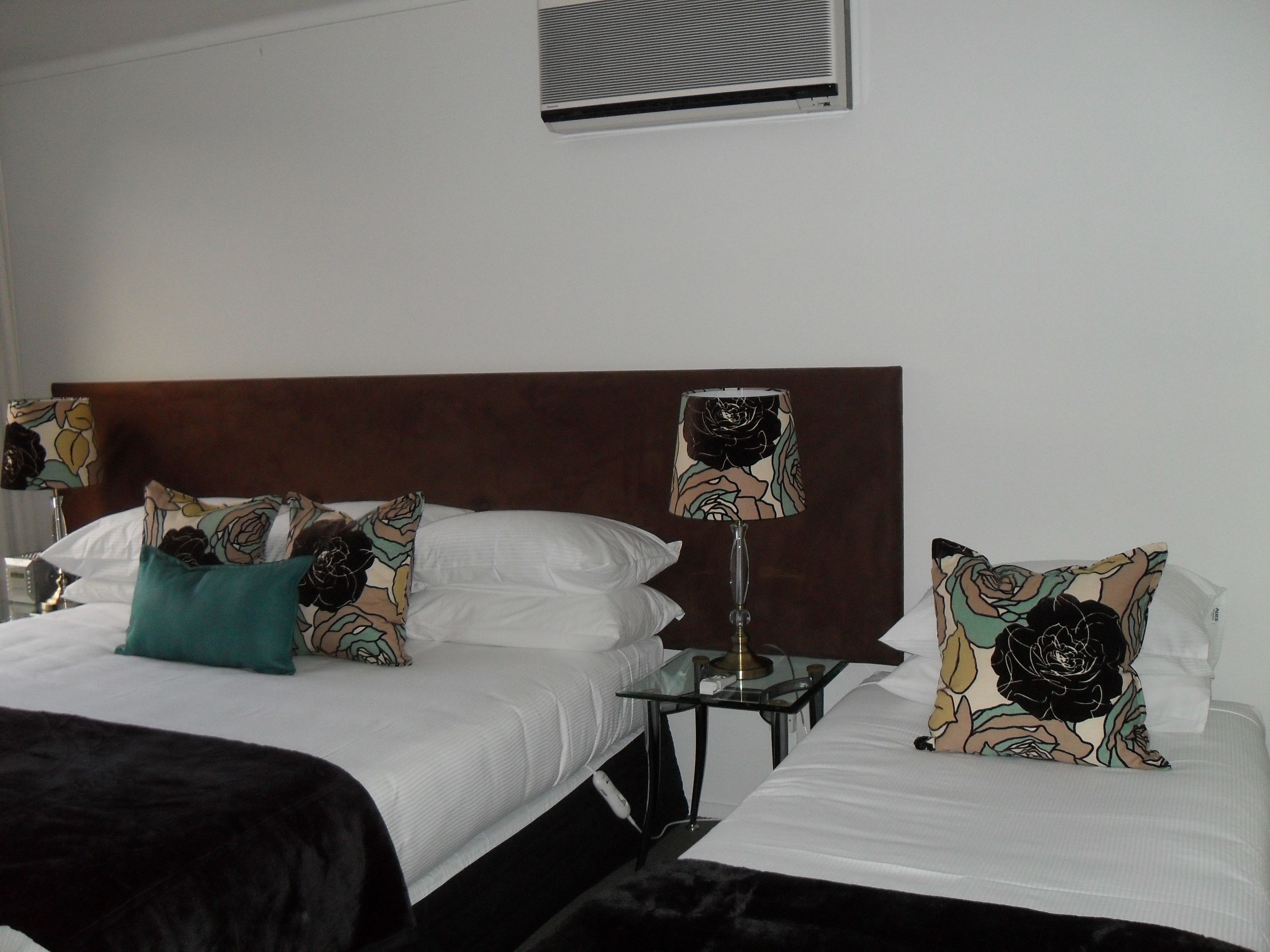 Habitación de hotel con dos camas, cada una con almohada estampada con motivos florales, una mesita de noche con lámpara y un respiradero de pared.