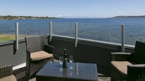 Een terras met rieten meubels, een tafel met twee wijnglazen en een fles, en uitzicht op een meer met bergen in de verte.