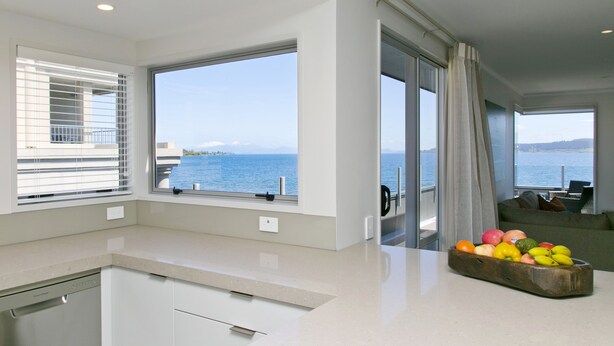 Een moderne keuken met een groot raam met uitzicht op zee, een fruitschaal op het aanrecht, en een zicht op de oceaan door het raam.