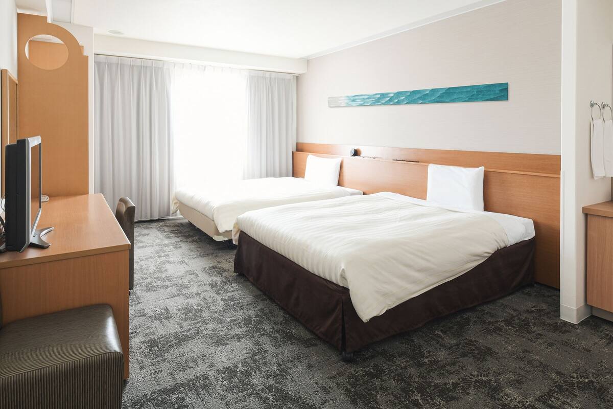 A hotel room with two beds, a desk with a computer, a chair, and a large window with curtains.